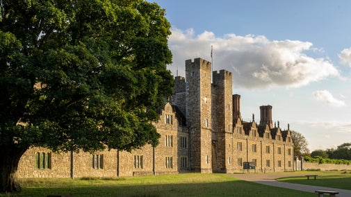 View of historic stone building, with blue sky and cloud above, green grass in the foreground and a large green-leaved tree to the left.